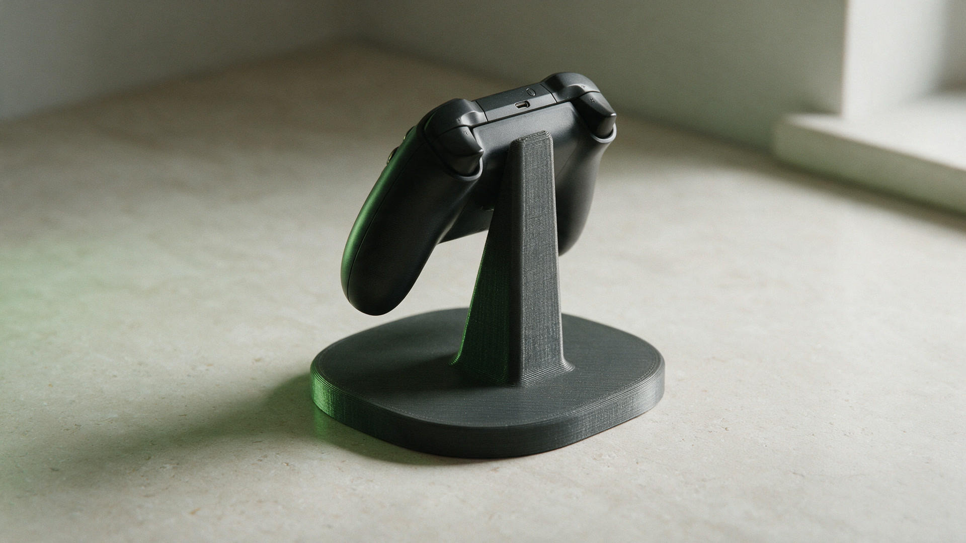 A charcoal printed controller stand holding a black game controller upright on a pale desk.