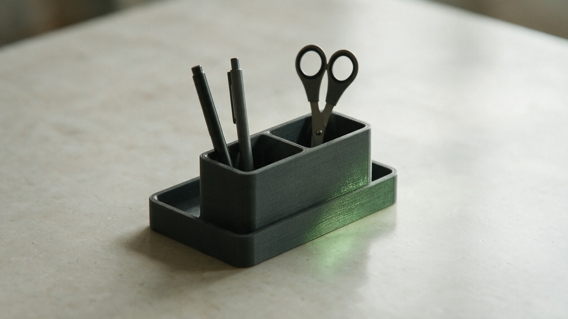A charcoal printed desk organiser tray holding two pens and a pair of scissors on a pale desk.