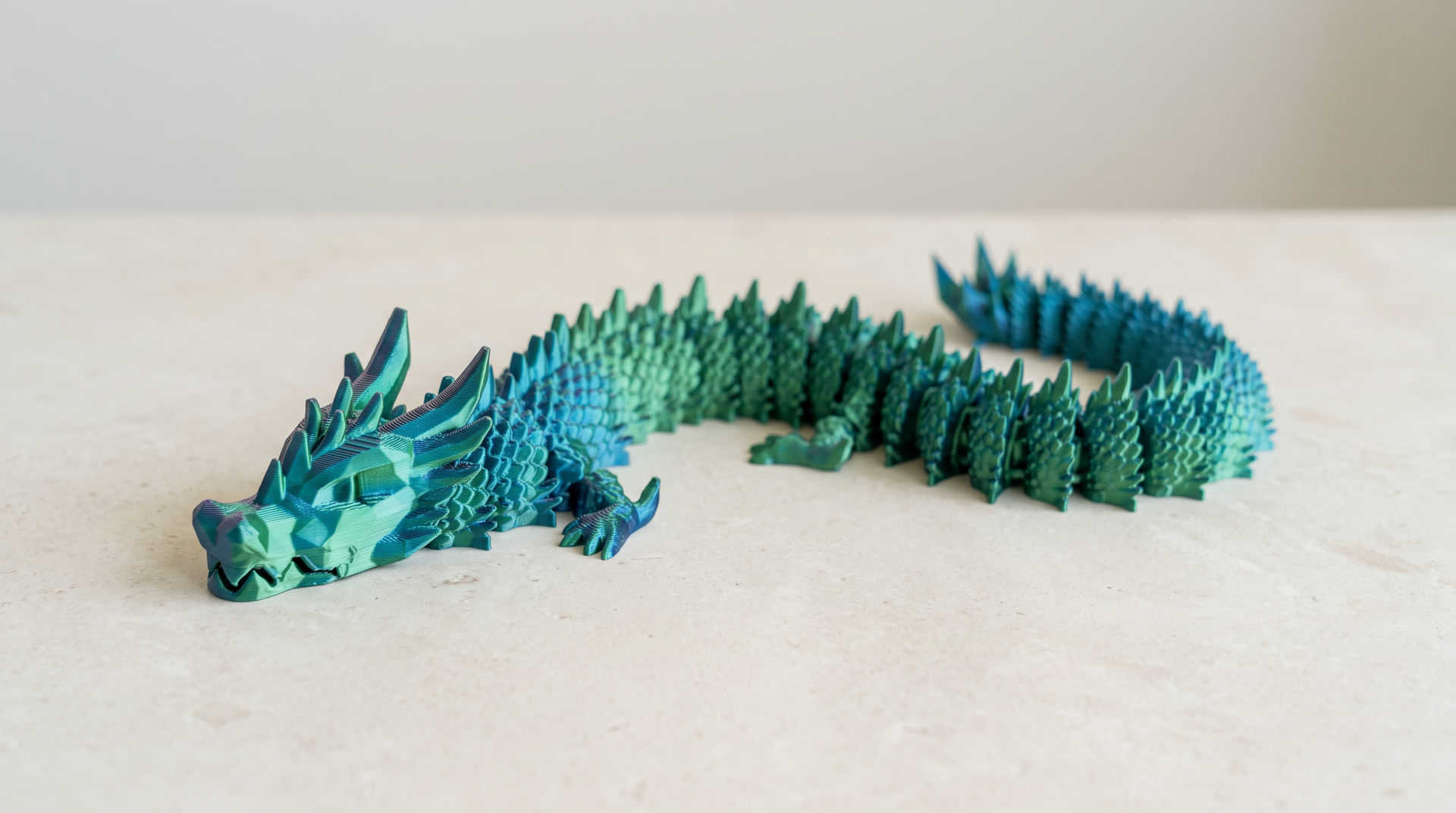 A long articulated printed crystal dragon in colour shifting teal and green, curving across a pale stone surface.
