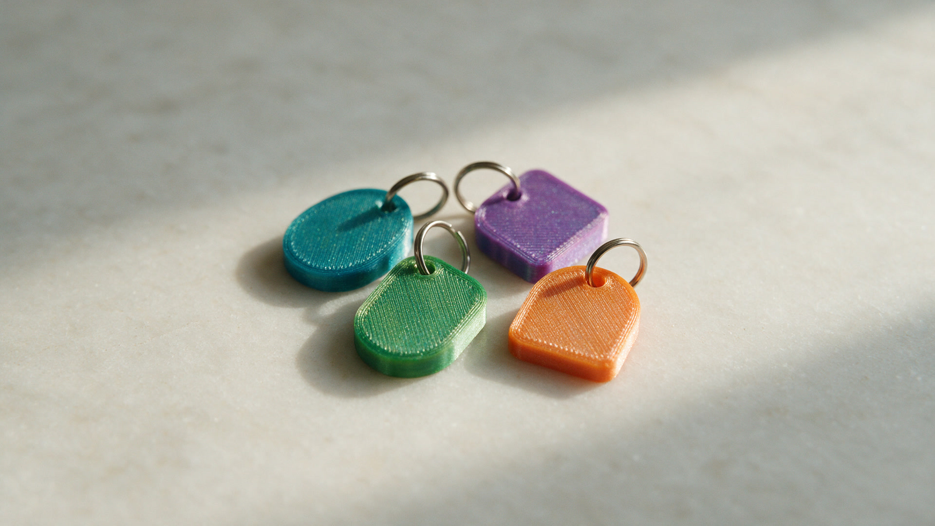 Four small printed keychain tags in teal, green, purple and orange on split rings, on a pale stone surface.