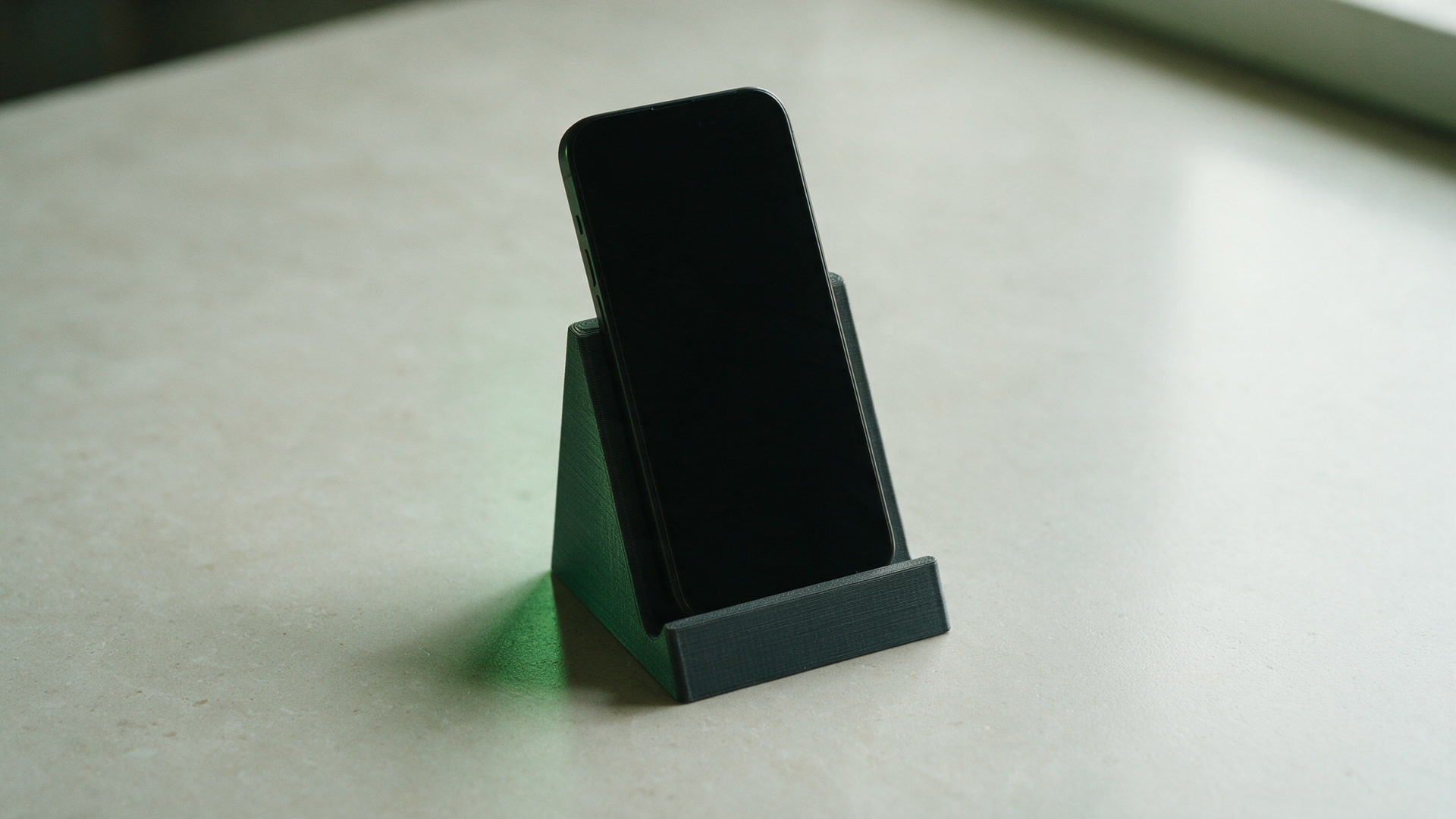 A charcoal printed angled phone stand holding a phone with a blank screen on a pale desk.