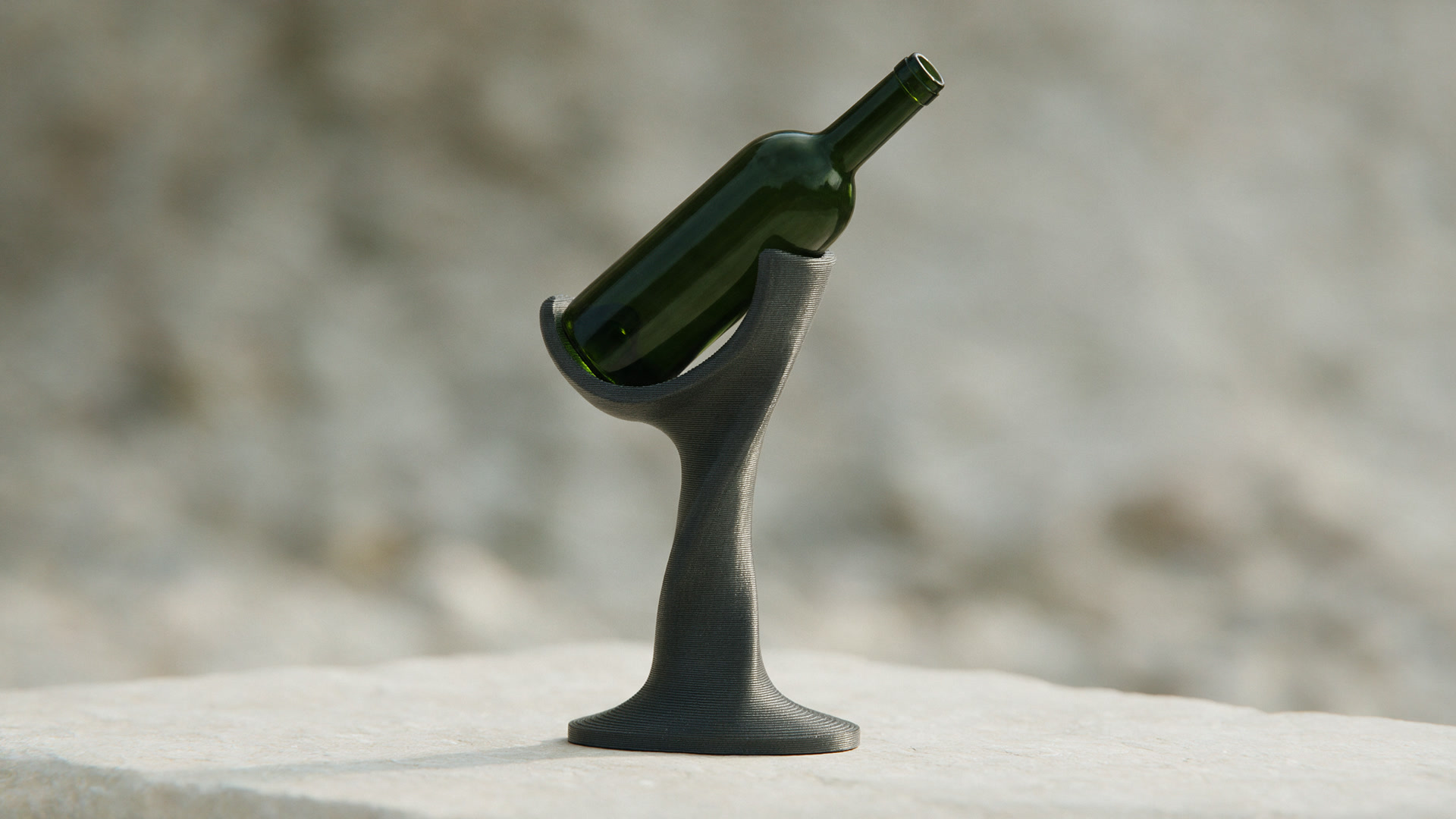 A sculptural charcoal printed wine holder balancing a dark green bottle on a pale stone surface.